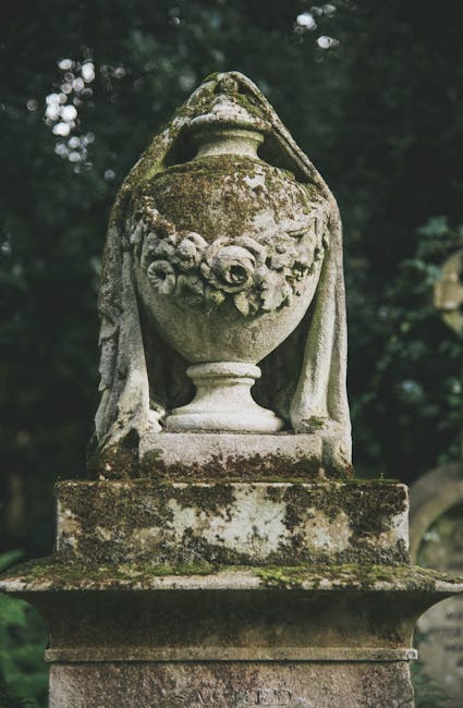 An old stone garden ornament shaped like an urn, featuring intricate carved floral patterns, sits atop a weathered stone pedestal. The urn has a rounded body with decorative garlands of flowers carved