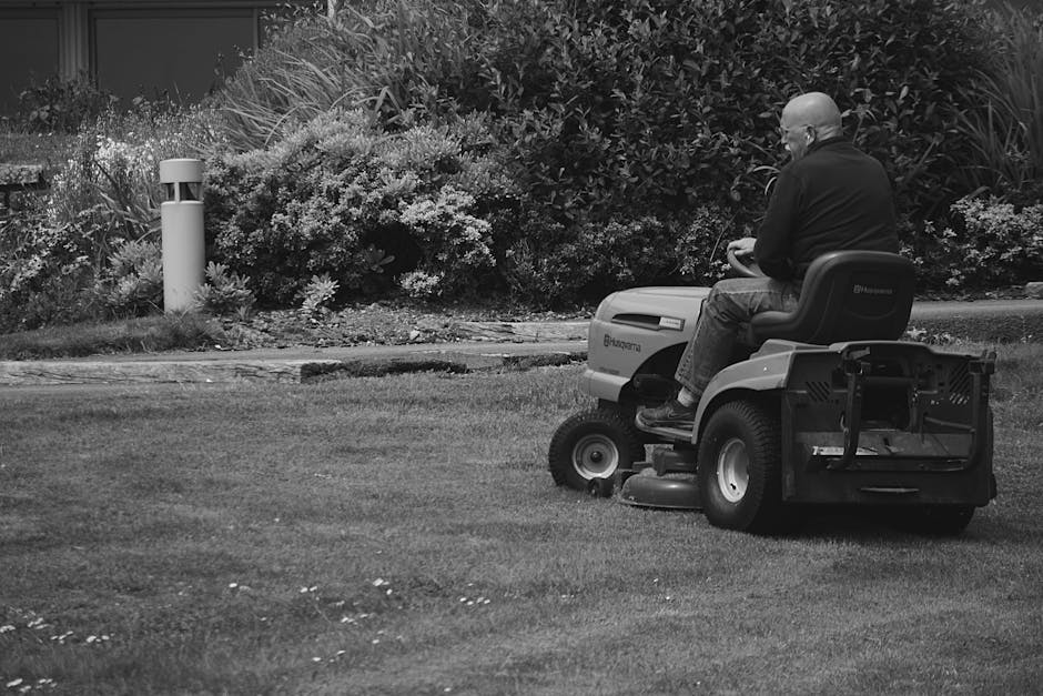 A person operating a ride-on lawn mower on a well-maintained grassy area of a garden. The lawn appears freshly cut, with visible mowing patterns creating a smooth texture. Behind the mower, a lush hed